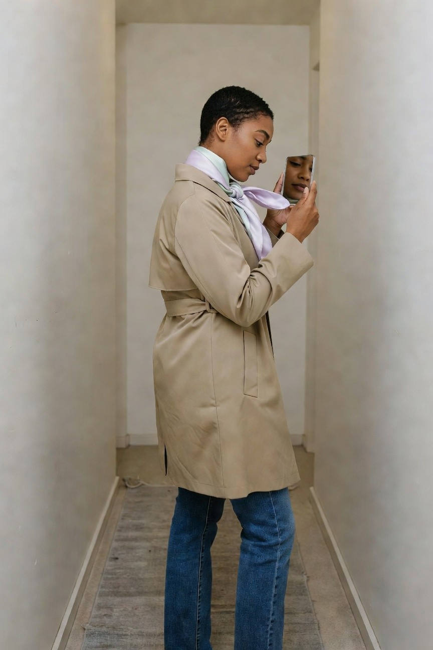 Woman in a beige belted trench coat over blue jeans with a pastel scarf tied at the neck holding a small mirror in a hallway.
