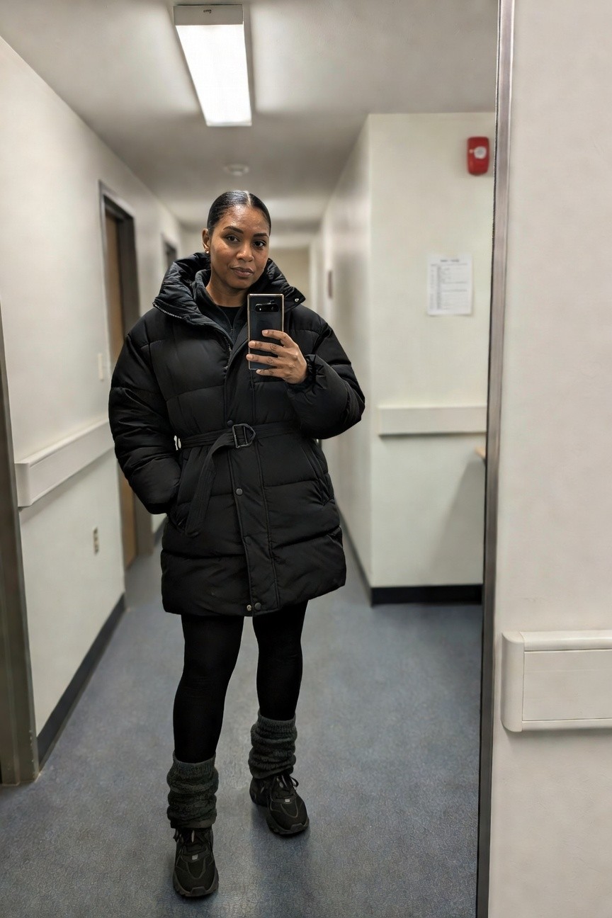 Woman wearing a long black puffer coat belted at the waist over black leggings with gray ankle cuffs and dark sneakers taking a mirror selfie in an indoor hallway