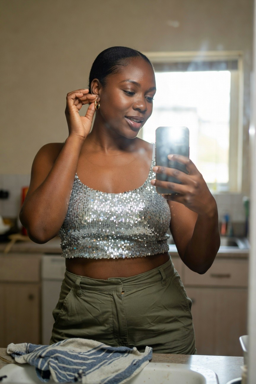 Woman in silver sequin crop top and high waisted olive pants taking a mirror selfie in a kitchen setting