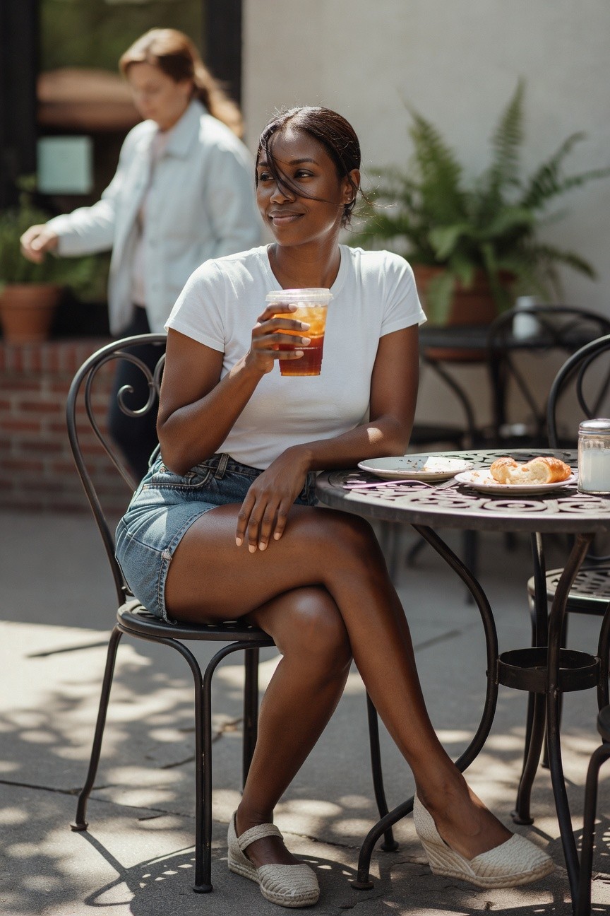 Black woman wearing a fitted white t-shirt tucked into a blue denim mini skirt with white espadrille wedges while seated at an outdoor cafe table holding an iced beverage.