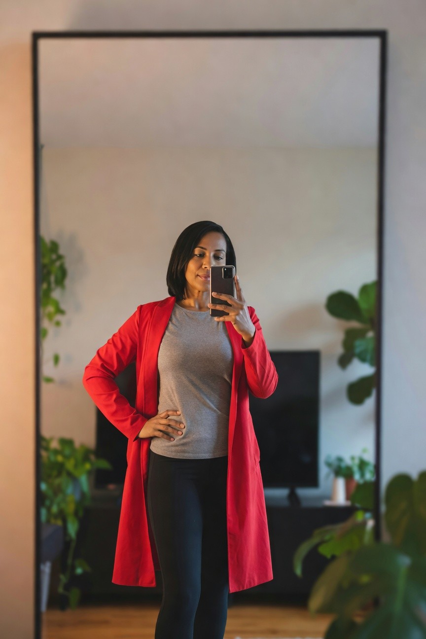 Woman taking a mirror selfie while wearing a long red coat over a grey t-shirt and black leggings.