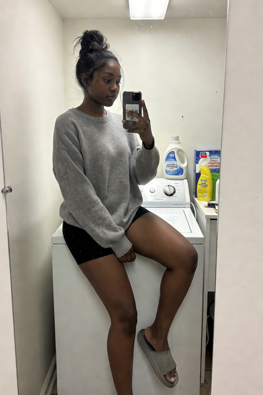Black woman in an oversized grey knit sweater and fitted black shorts with grey slides taking a mirror selfie while seated on a white washing machine.