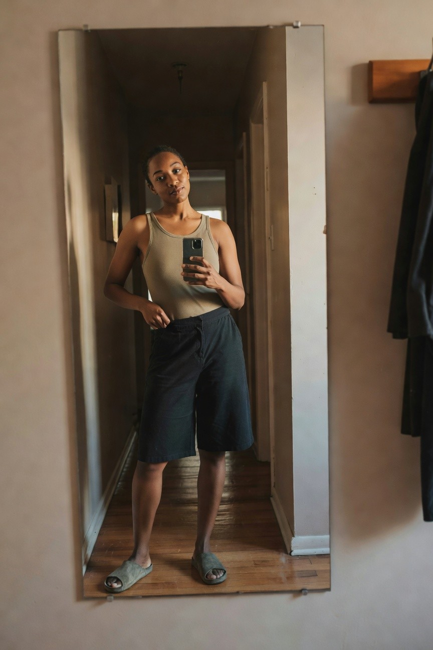 Woman taking a mirror selfie in a beige tank top, navy knee-length drawstring shorts, and gray slide sandals.