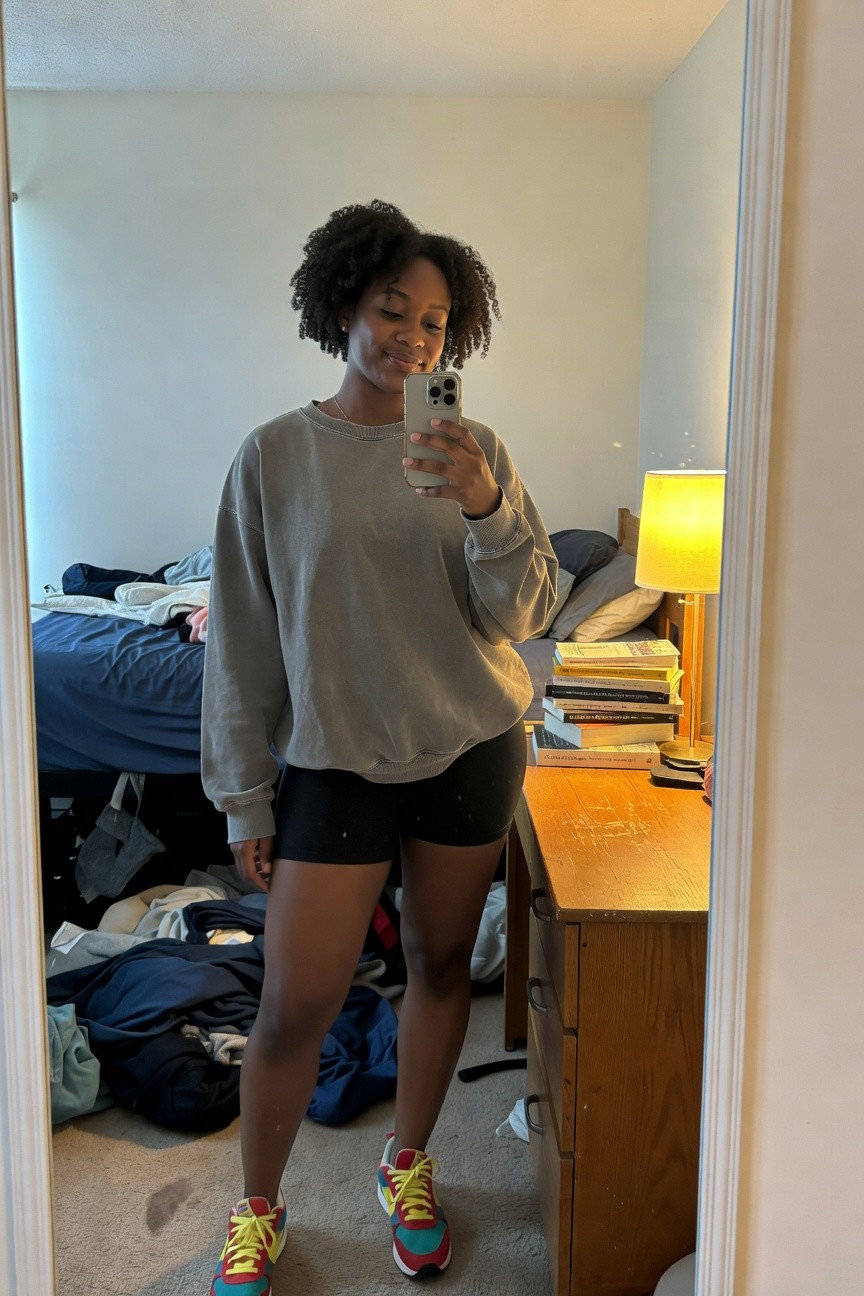 A Black woman in an oversized grey sweatshirt, black athletic shorts, and multicolored red yellow teal sneakers takes a mirror selfie in a bedroom setting.