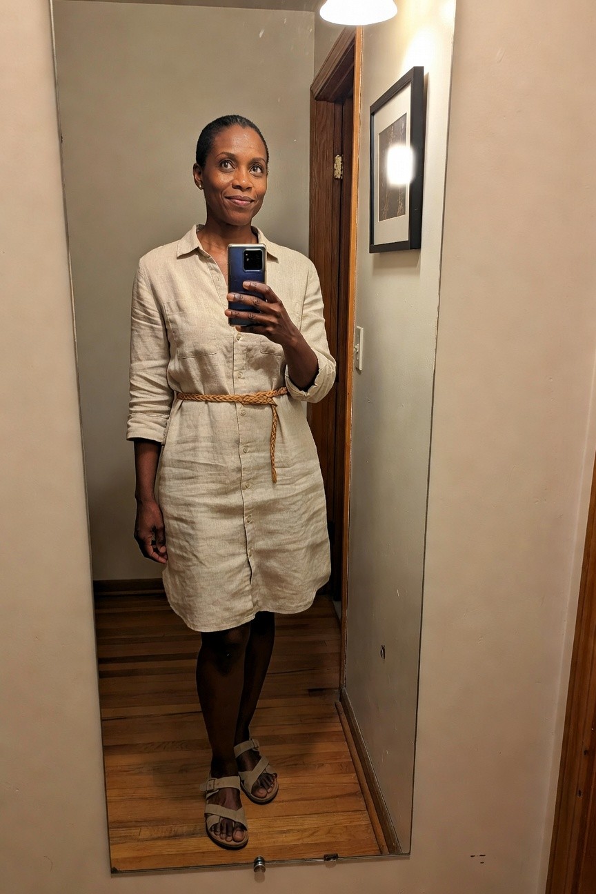 Woman wearing a beige linen button front shirt dress with a natural braided belt black tights and beige sandals holding a phone for a mirror selfie in a hallway.