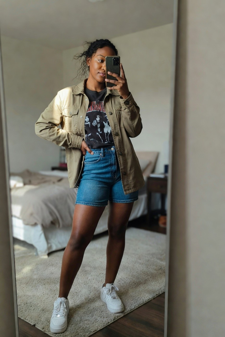 Woman in mirror selfie wearing khaki utility jacket over black graphic band tee tucked into blue denim shorts paired with white sneakers.
