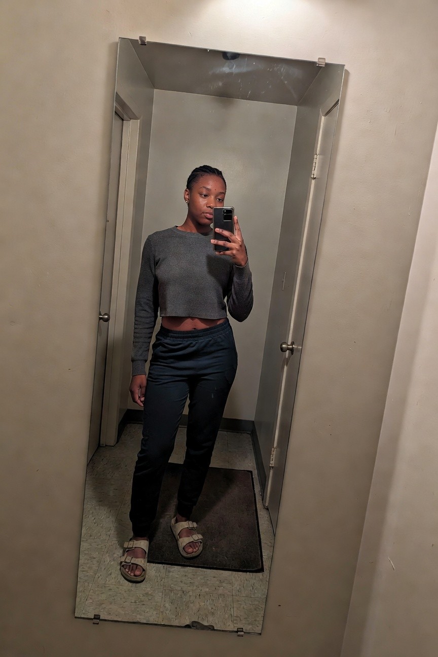 A young Black woman taking a mirror selfie wearing a gray cropped long-sleeve sweater, dark blue sweatpants, and beige sandals.