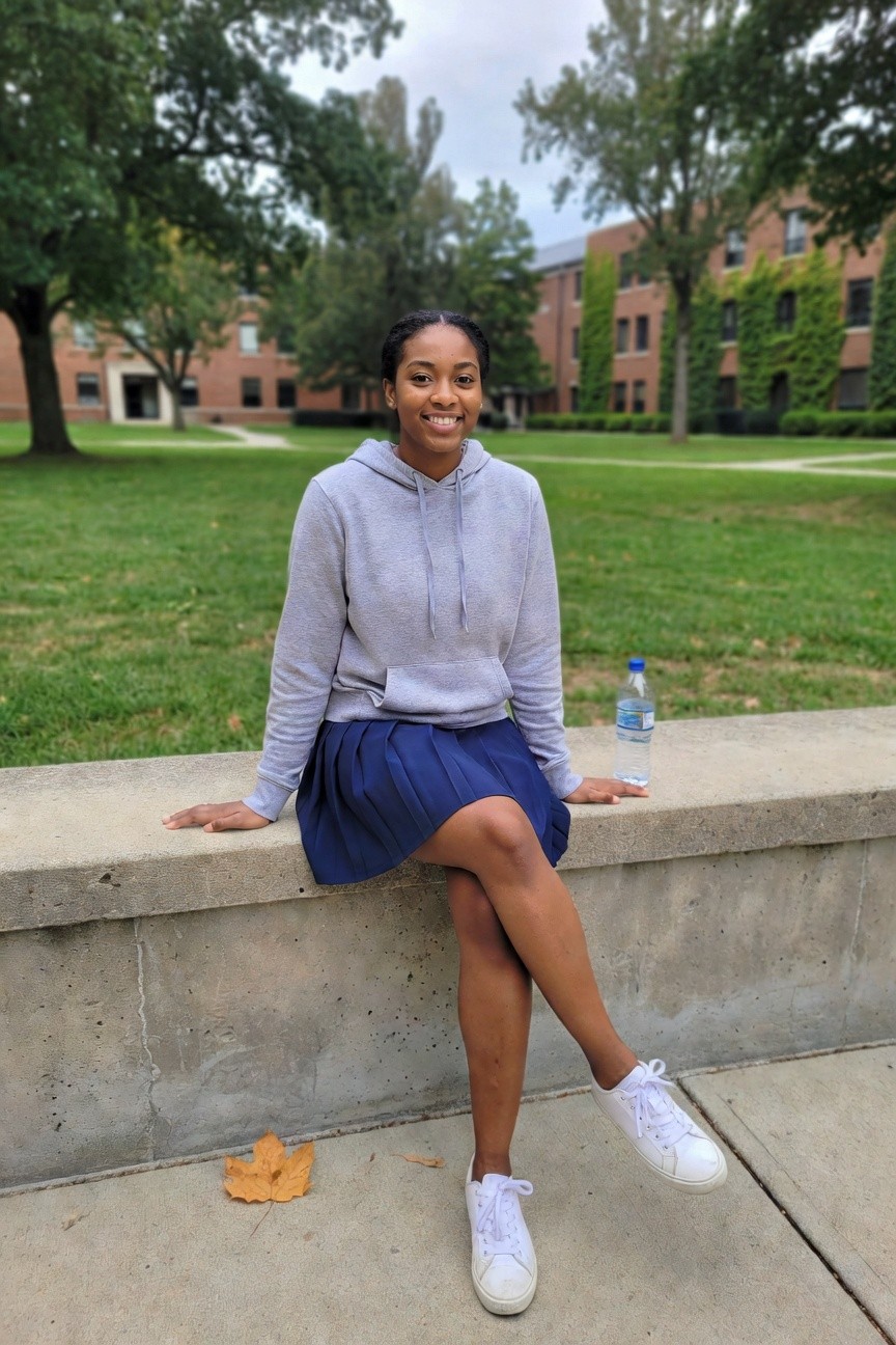 woman in light gray hoodie and blue pleated skirt with white sneakers sitting on a concrete ledge outdoors