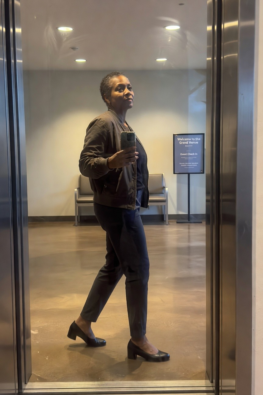 Woman in brown bomber jacket over dark top with slim dark pants and black block heel shoes standing indoors holding a phone.