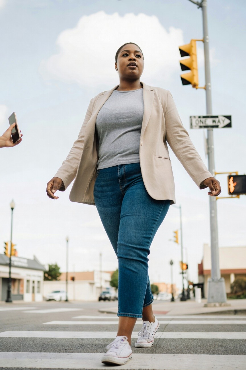 Plus size Black woman in a beige blazer over a gray t-shirt paired with cropped blue jeans and white sneakers while crossing a street
