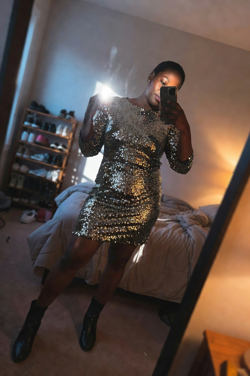 Woman wearing a short silver sequin mini dress with three-quarter sleeves and black ankle boots while taking a mirror selfie indoors