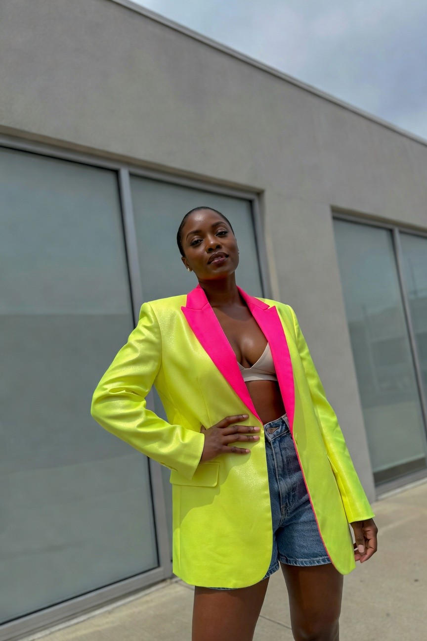 Black woman in an oversized neon yellow blazer with pink lapels worn open over a beige crop top and paired with blue denim shorts standing in front of a building.