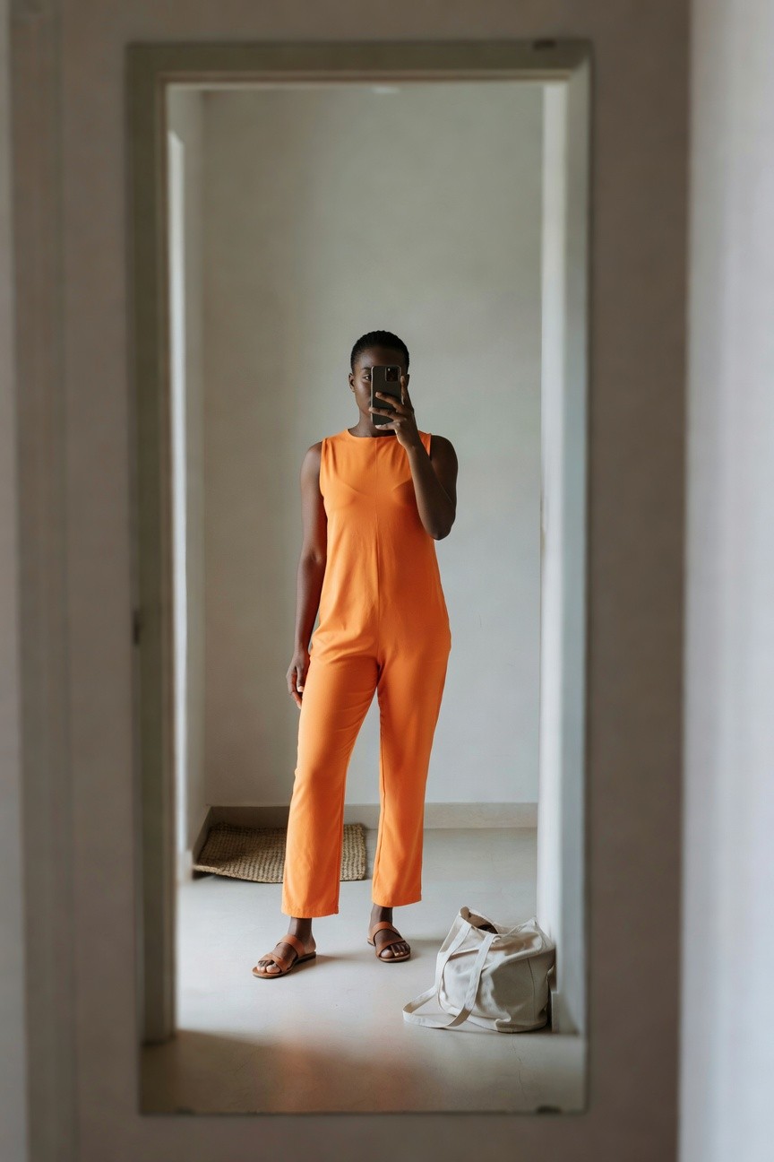 A Black woman taking a mirror selfie while wearing a bright orange sleeveless jumpsuit with cropped straight legs and brown slide sandals.