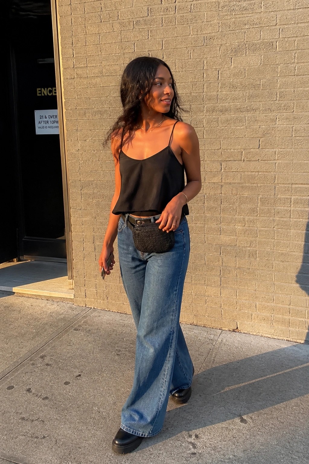 Woman in black camisole and wide-leg blue jeans with fanny pack by brick wall