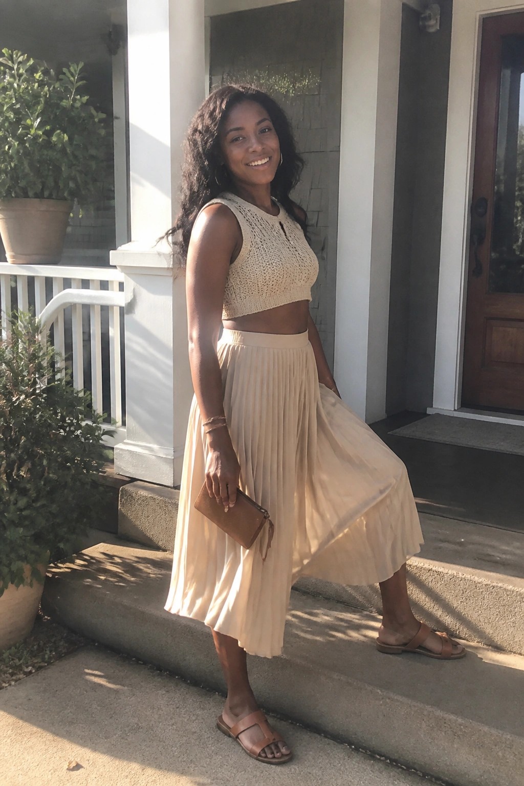 Woman in beige crochet crop top and pleated skirt on sunny porch steps