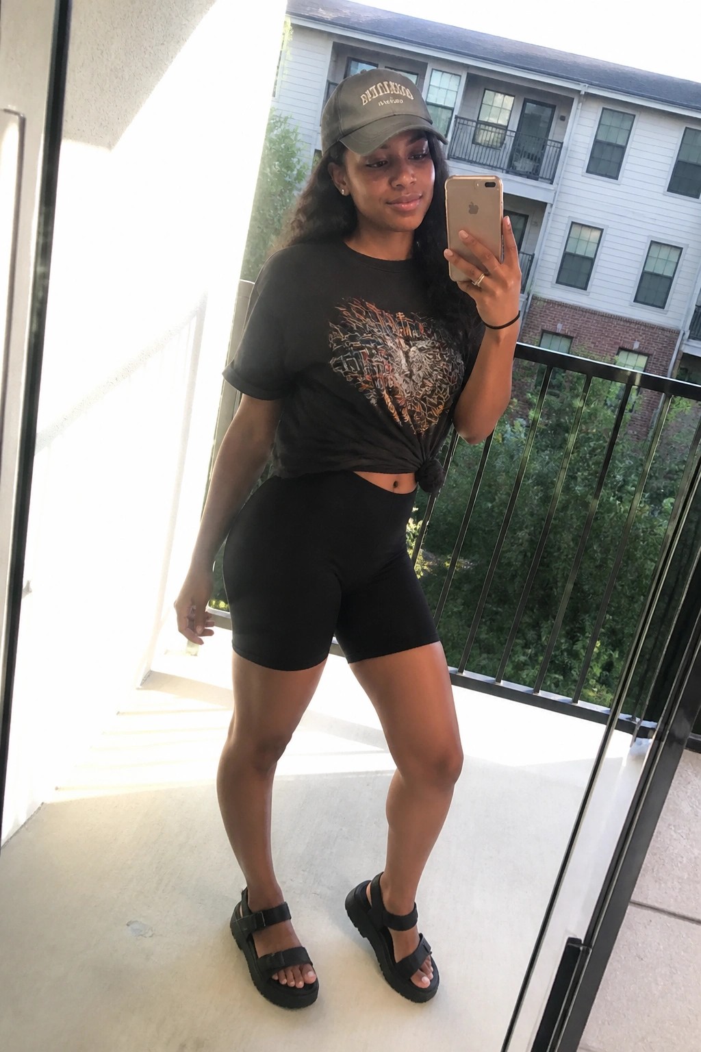 Woman in green cap, graphic tee, and biker shorts taking balcony selfie with phone