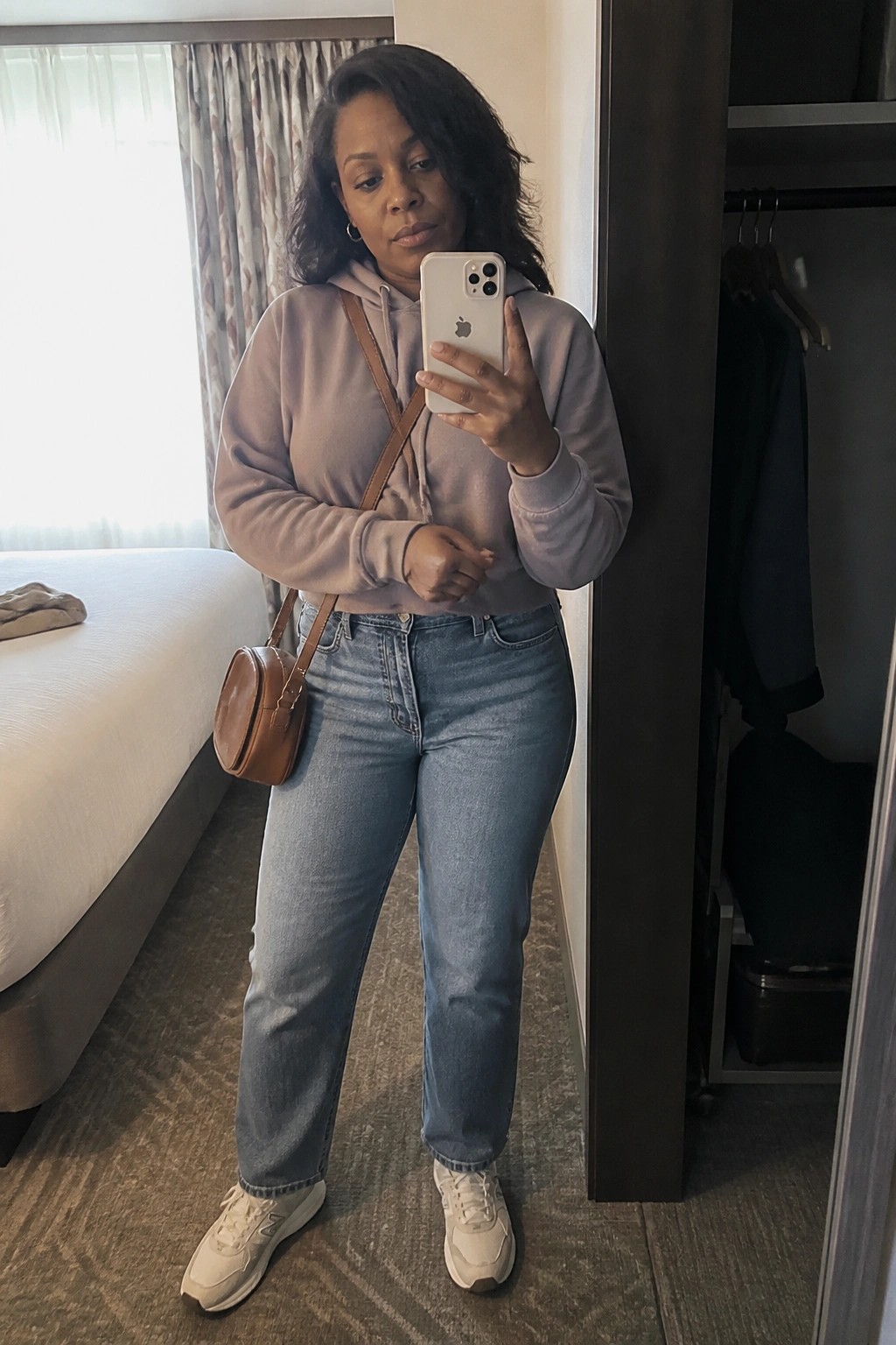 Woman wearing a cropped lavender hoodie, blue straight-leg jeans, white sneakers, and a brown leather crossbody bag while taking a mirror selfie.