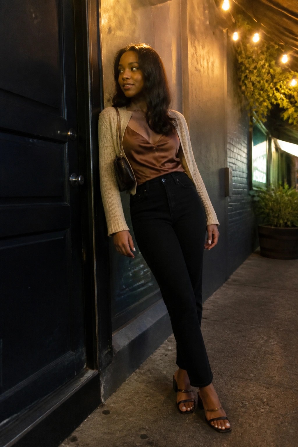 Woman in brown satin top, beige cardigan, and black jeans leaning on wall at night