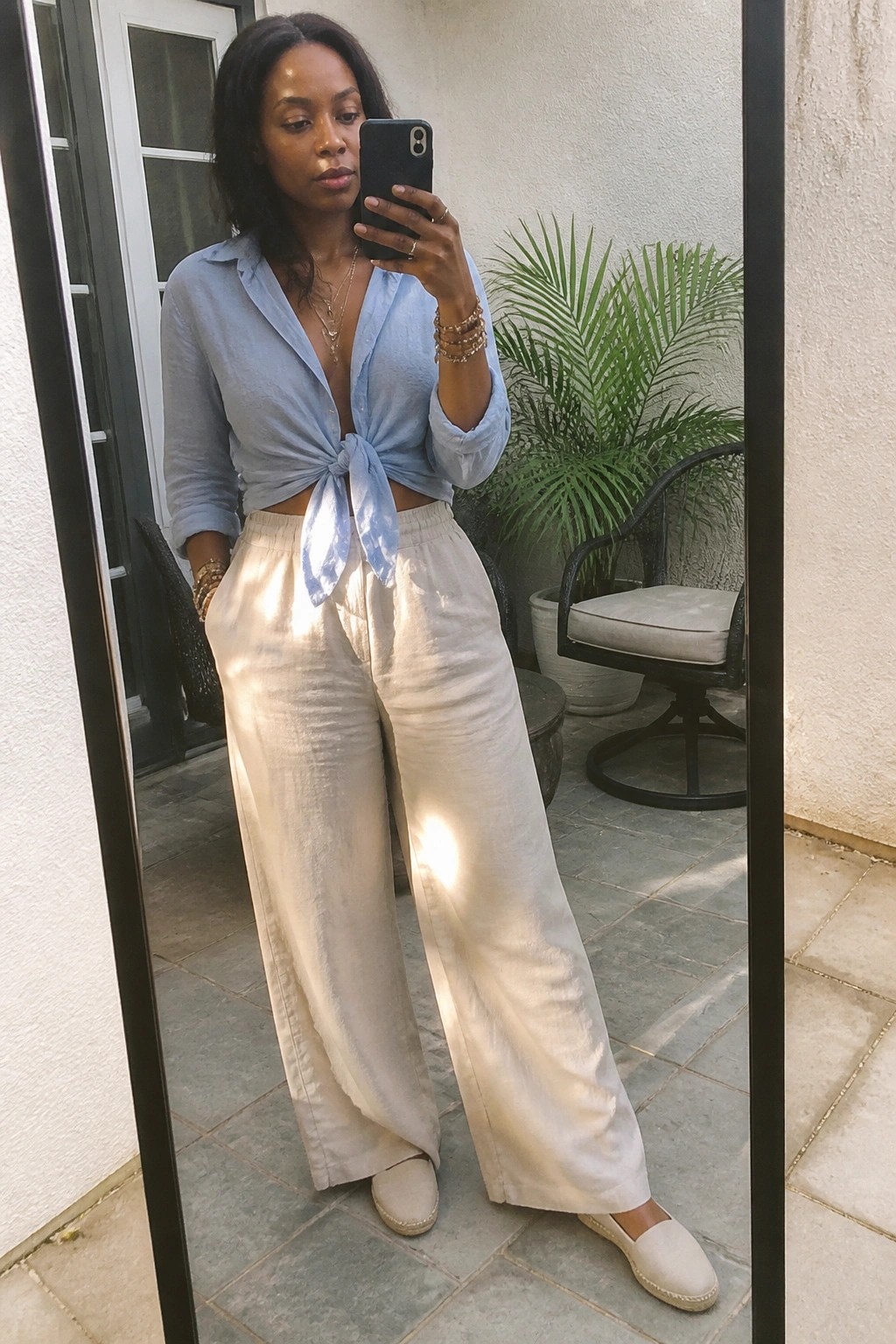 Woman in light blue tied shirt and cream wide-leg pants taking mirror selfie outdoors.