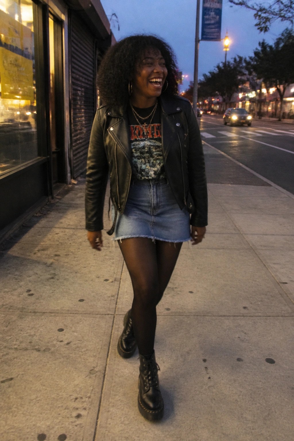 A woman wearing a black leather jacket over a graphic KISS t-shirt, paired with a blue denim mini skirt, black tights, and black combat boots, accessorized with layered necklaces and hoop earrings.