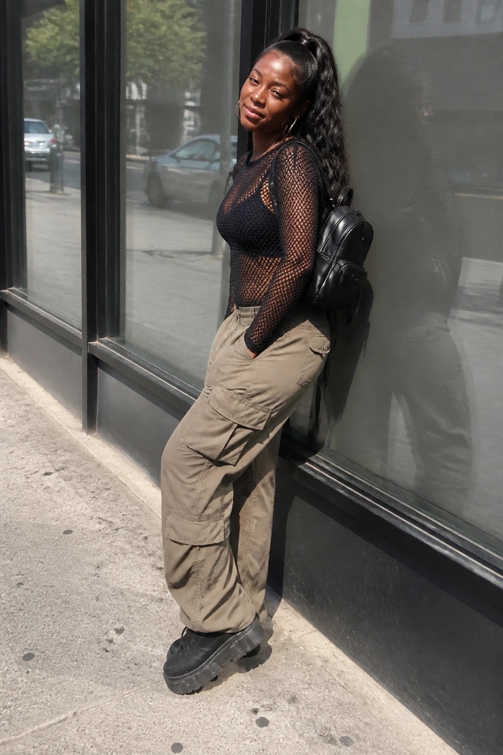 Woman in black mesh top and cargo pants leaning against glass storefront with backpack
