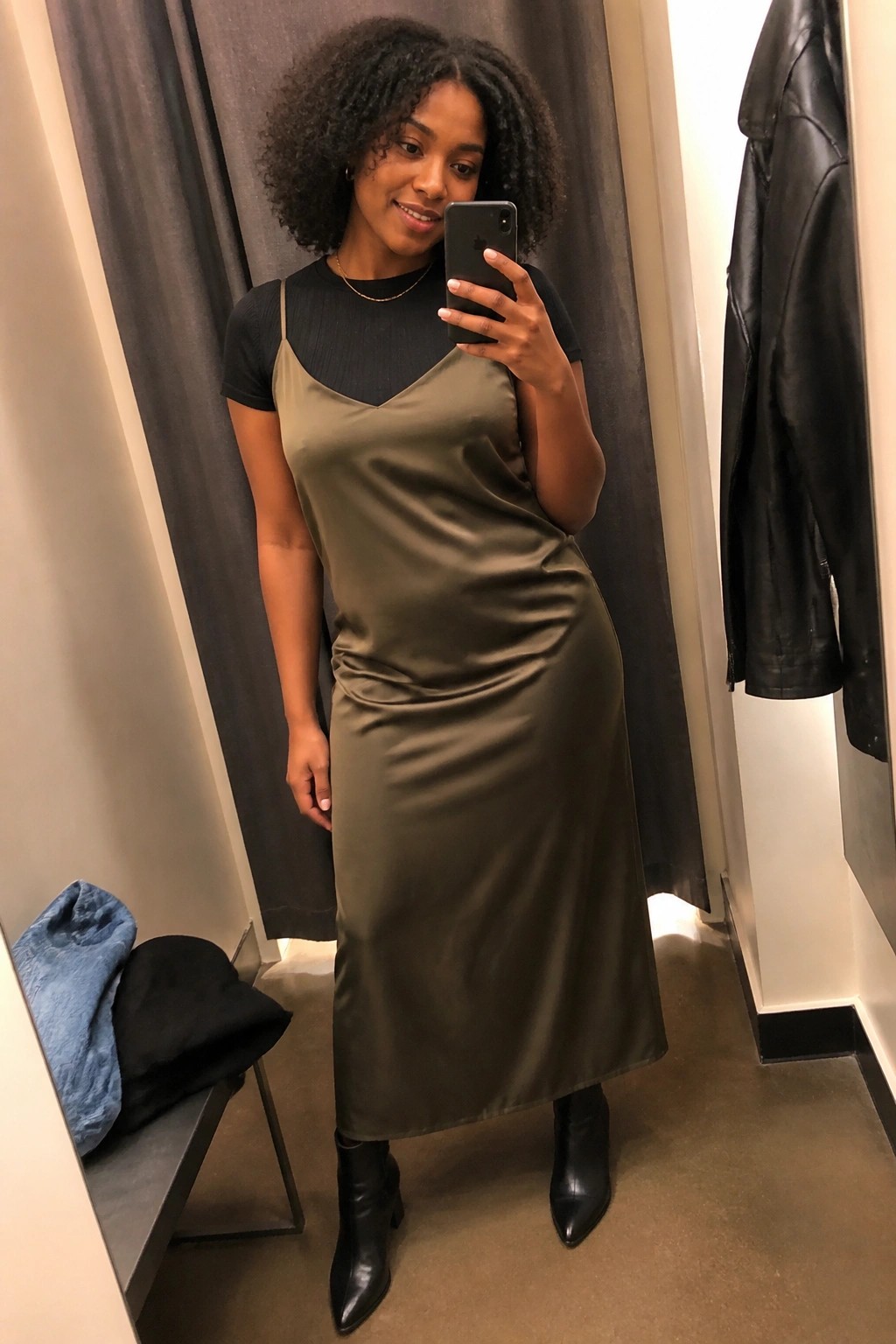 Woman in olive satin slip dress taking mirror selfie in fitting room.