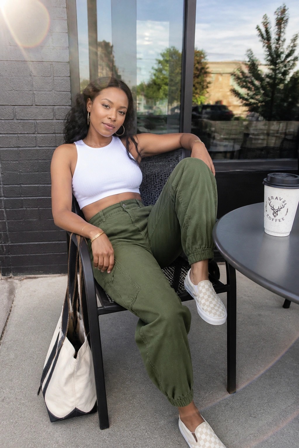 Woman in white crop top and green cargo pants seated at outdoor cafe with coffee and tote bag