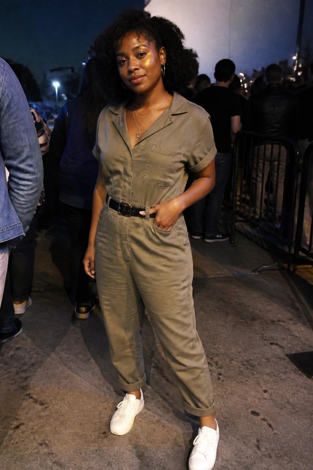 Woman in olive jumpsuit with black belt and white sneakers at night event