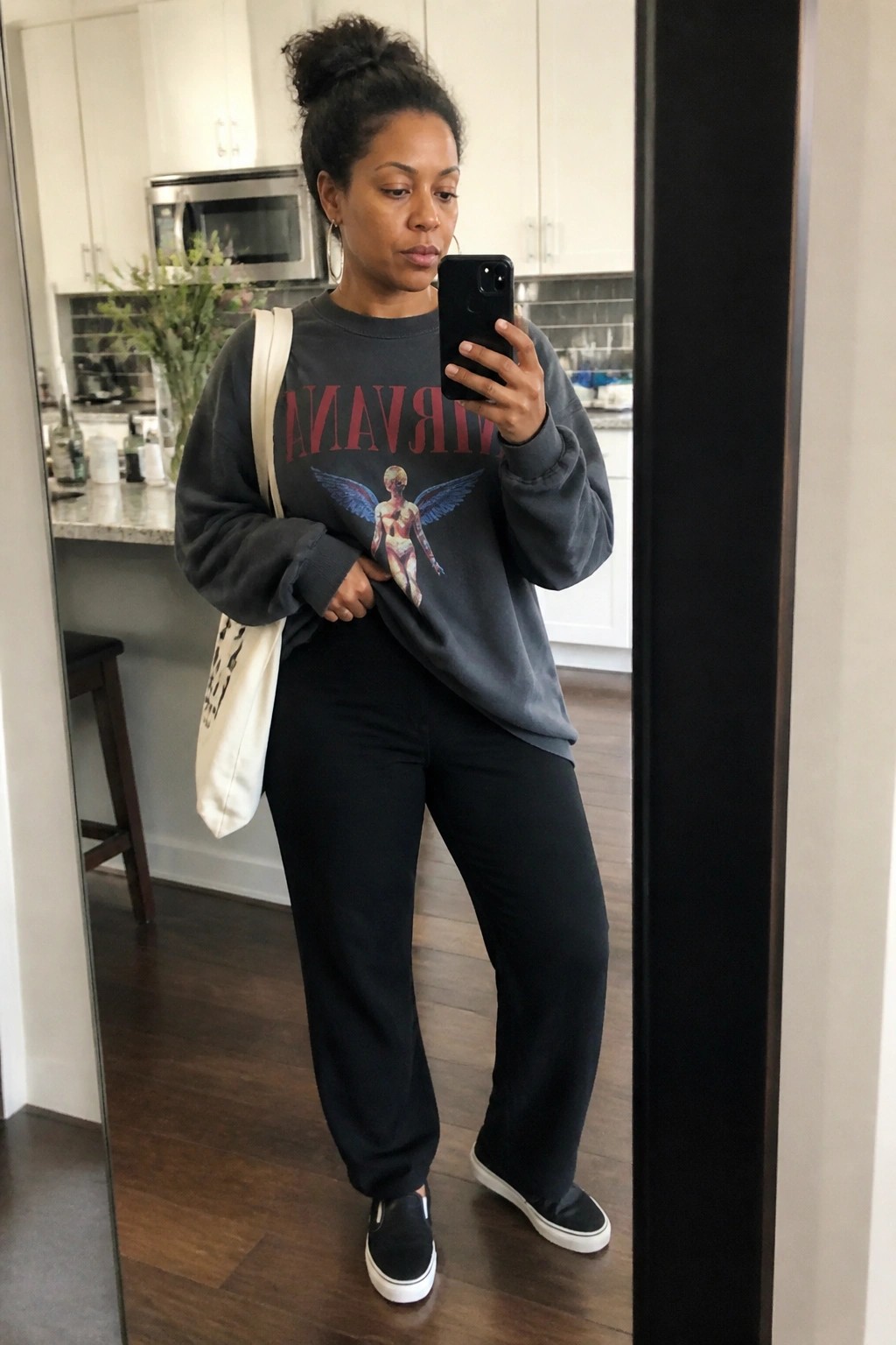 Woman wearing an oversized gray Nirvana graphic sweatshirt tucked slightly at the front, black straight-leg pants, black slip-on sneakers, and carrying a white tote bag while taking a mirror selfie.