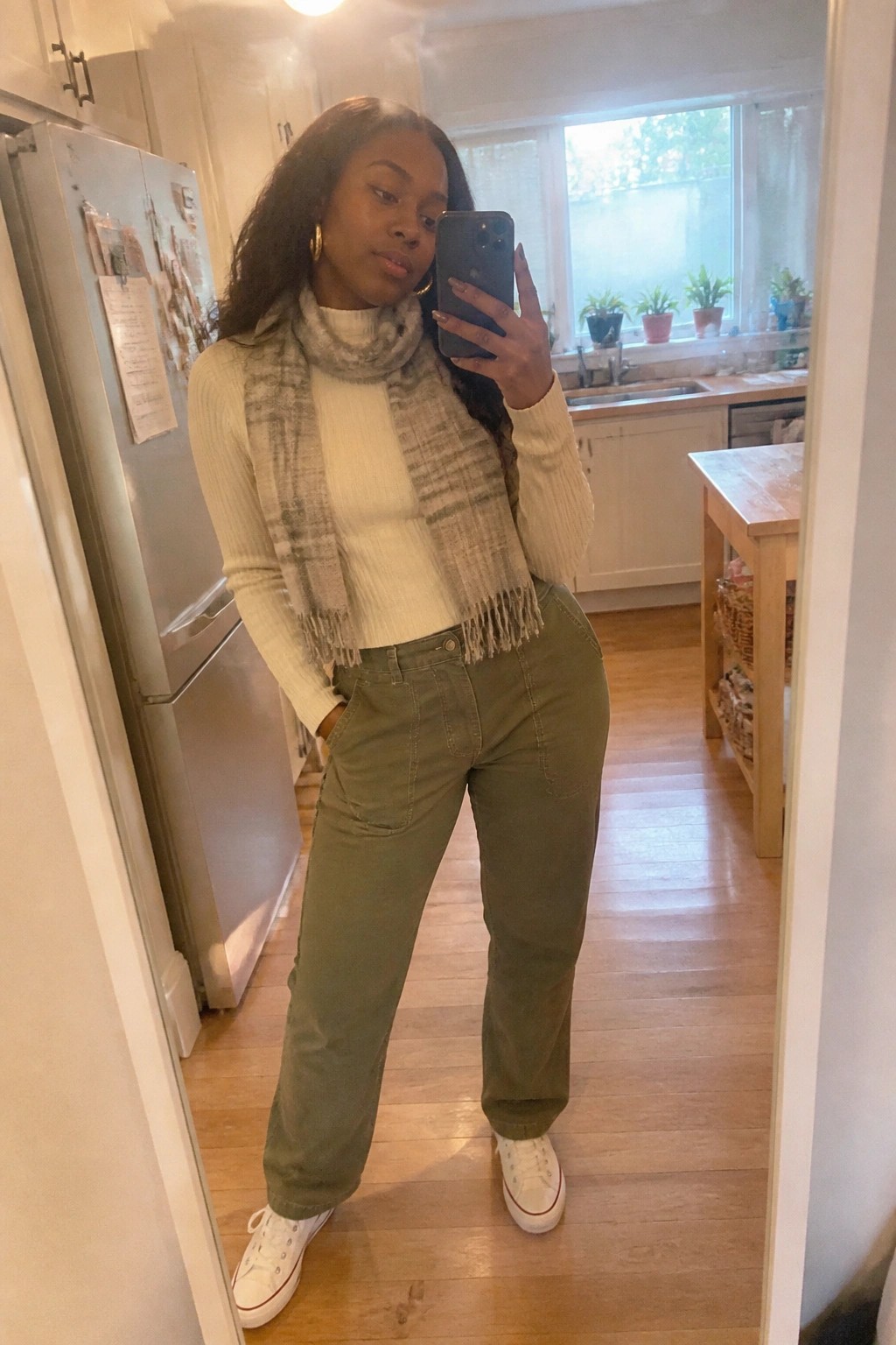 Woman in white sweater and plaid scarf taking mirror selfie in kitchen