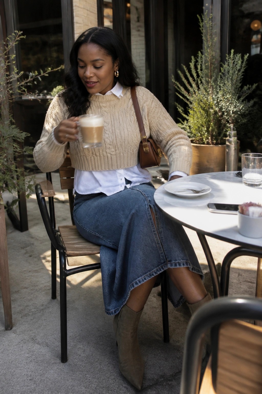 Woman wearing a cropped beige cable knit sweater layered over a white button down shirt, paired with a blue denim midi skirt and taupe suede ankle boots, holding a coffee cup at an outdoor table.