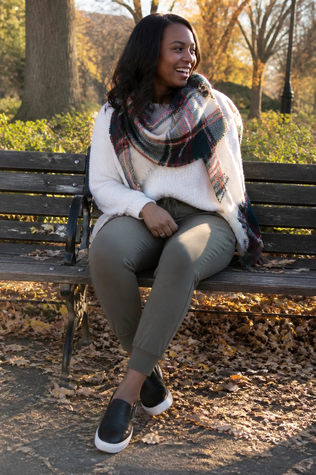 Smiling woman in white sweater and plaid scarf on autumn park bench with fallen leaves