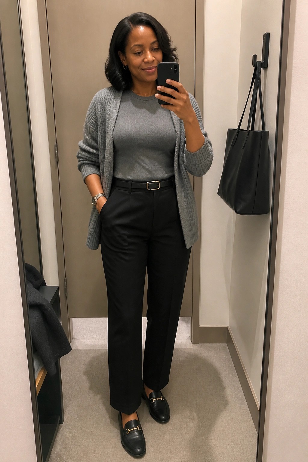Woman taking mirror selfie in gray cardigan, gray top, and black pants in dressing room