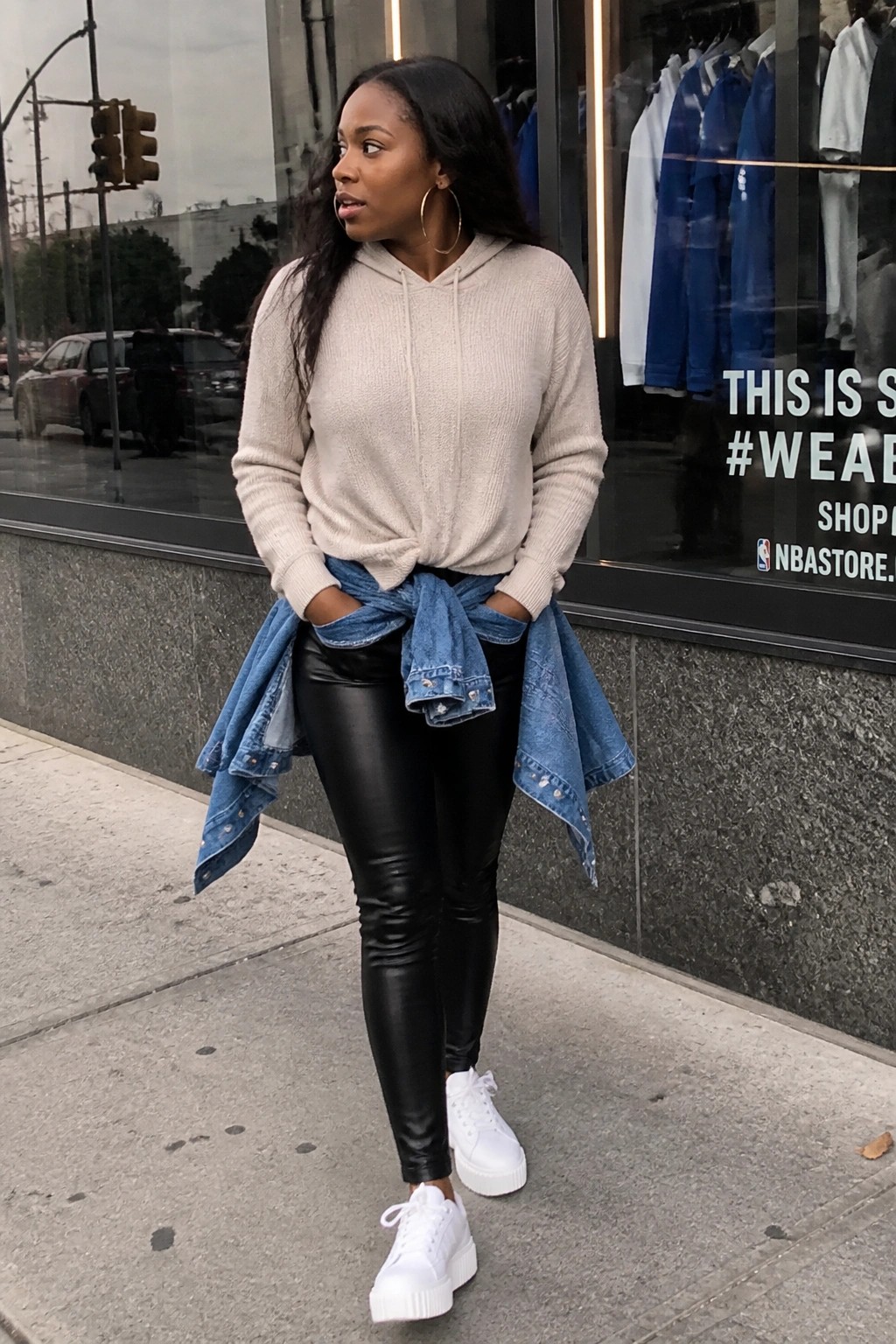 Woman in beige hoodie, black leggings, tied denim jacket, and white sneakers on sidewalk
