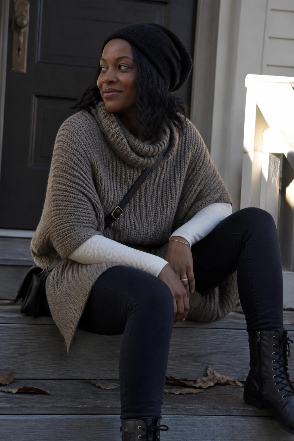 Woman wearing a taupe chunky knit poncho sweater over a white long sleeve top with black jeans, black lace up boots, and a black beanie, seated on wooden steps.