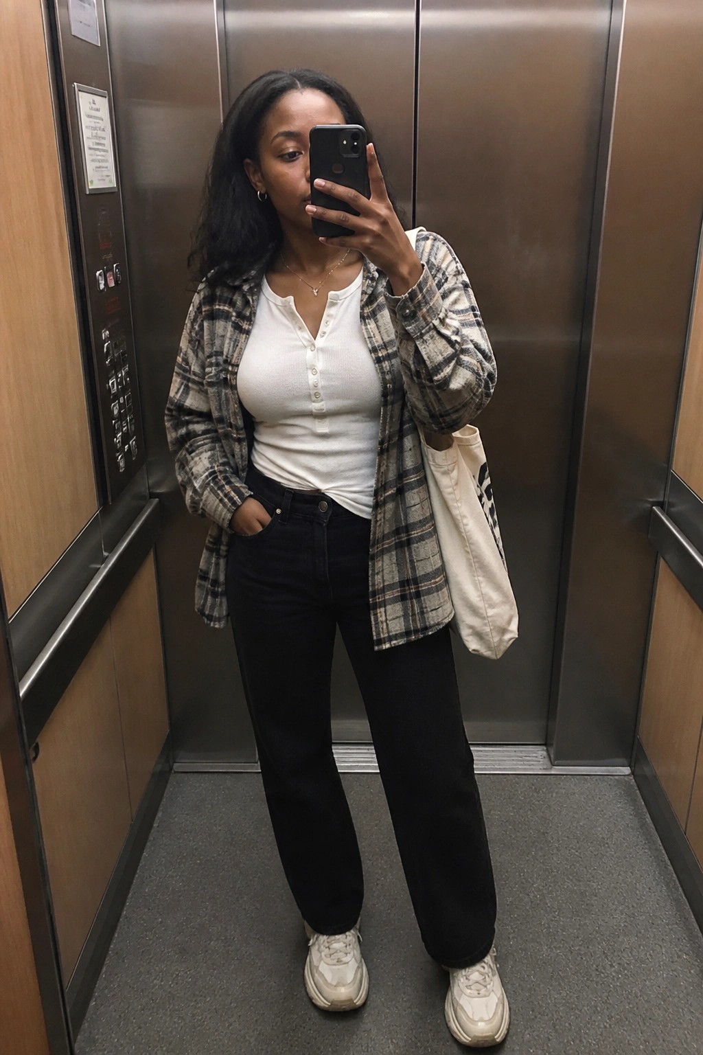 Woman standing in an elevator wearing an open plaid flannel shirt over a white ribbed henley, dark jeans, white chunky sneakers, layered gold necklaces, and carrying a canvas tote bag.