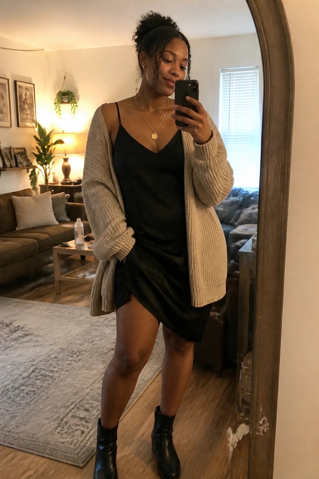 Woman wearing a black satin slip dress layered under an oversized beige chunky knit cardigan with black ankle boots and layered gold necklaces.