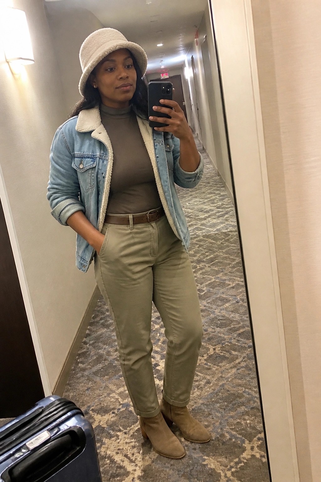 Woman in sherpa denim jacket and green pants taking mirror selfie in hallway.