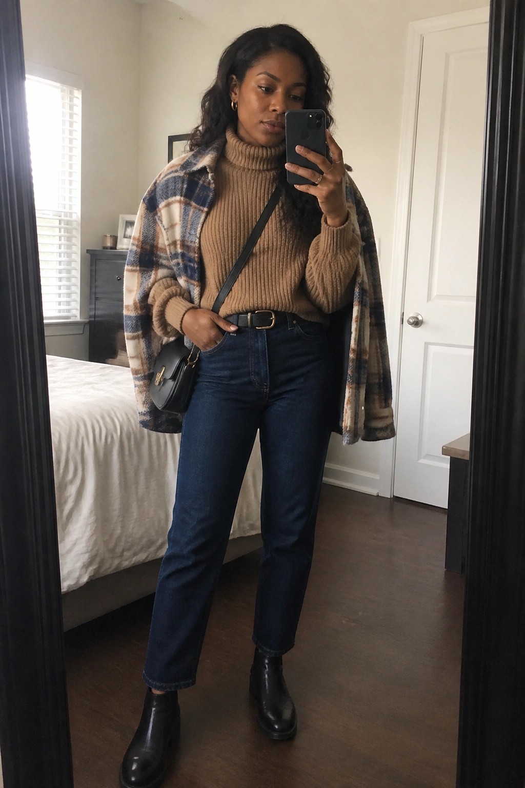 Woman in plaid shacket over tan turtleneck and jeans taking bedroom mirror selfie.