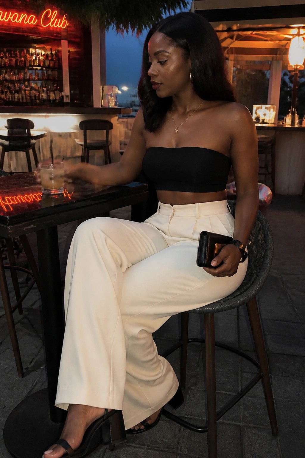 Woman in black strapless top and white pants at outdoor bar holding drink and clutch