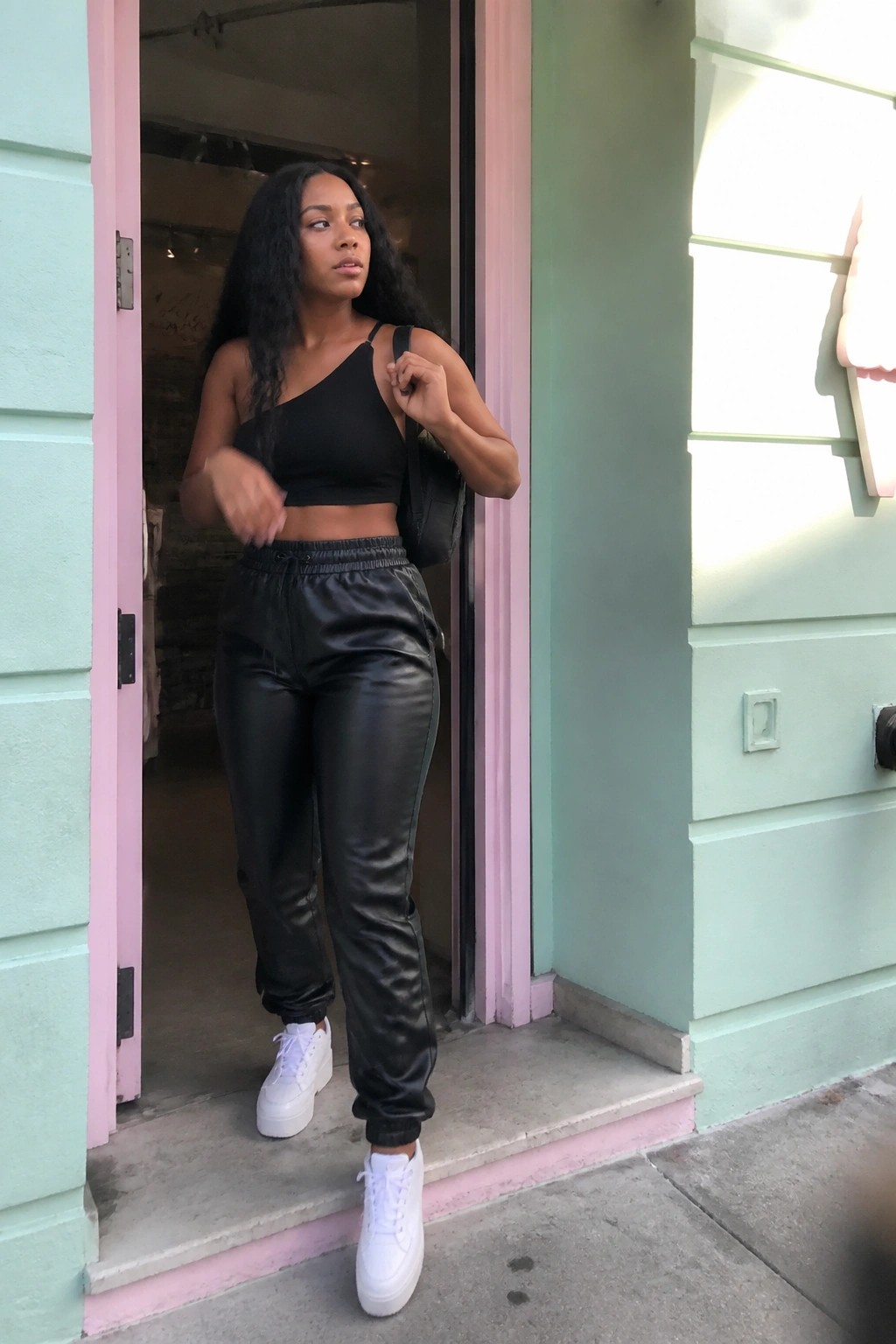 Woman in black one-shoulder crop top, black faux leather jogger pants, and white platform sneakers standing in a doorway.