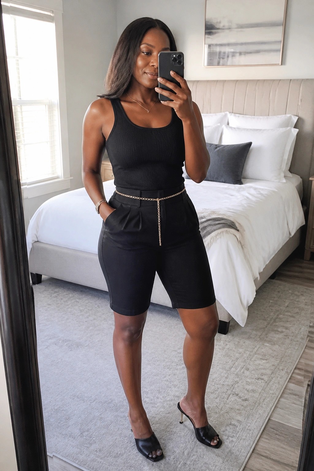 Woman wearing a black ribbed tank top, black high-waisted Bermuda shorts with gold chain belt, and black peep-toe heeled mules while taking a mirror selfie in a bedroom.