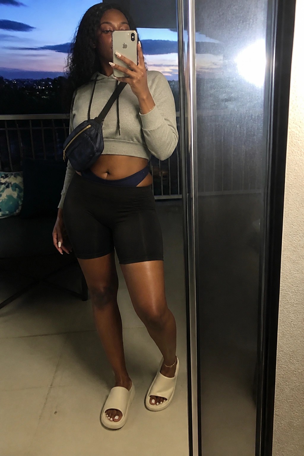 Woman taking mirror selfie on balcony in gray cropped hoodie and black shorts.