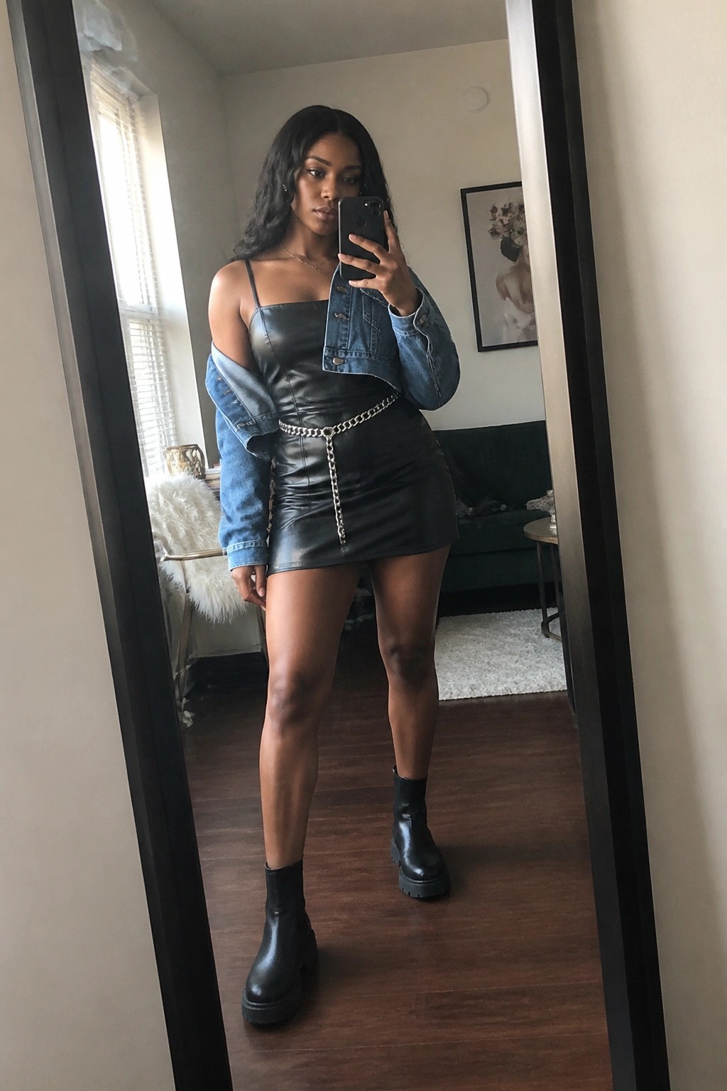 Woman in black leather mini dress with off-shoulder blue denim jacket, silver chain belt, and black platform ankle boots taking a mirror selfie.