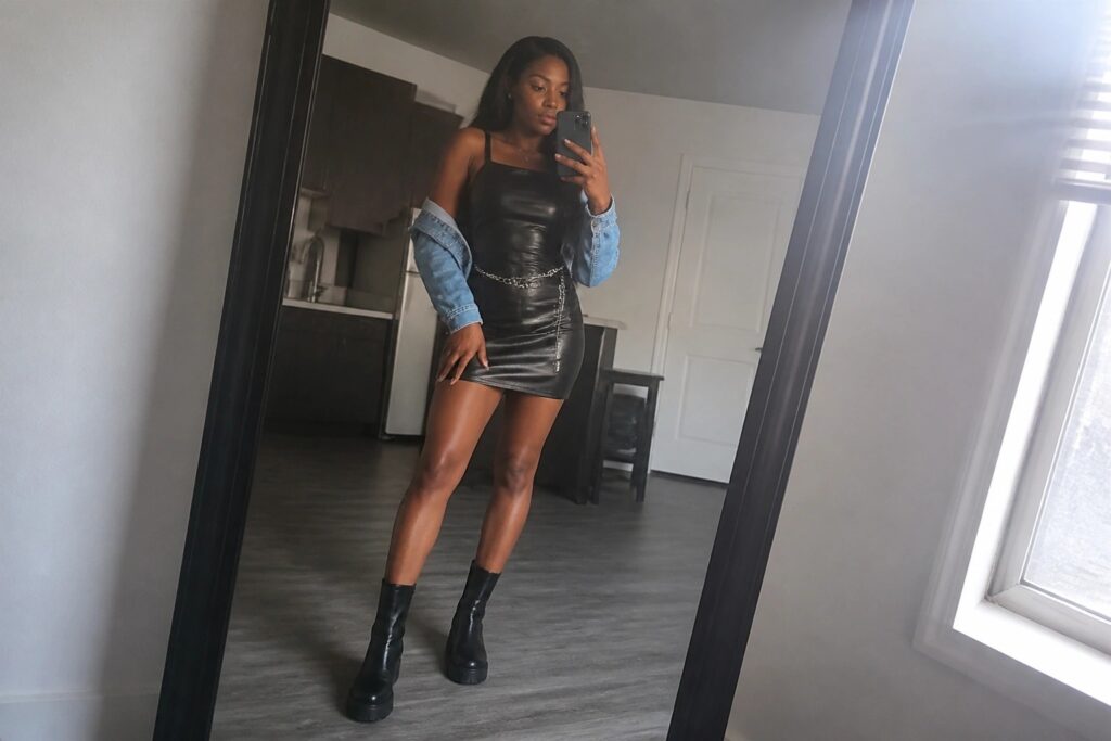 18 Edgy Miami Outfits for Black Women That Go From Beach to Nightlife Woman in black leather mini dress with off-shoulder blue denim jacket, silver chain belt, and black platform ankle boots taking a mirror selfie.