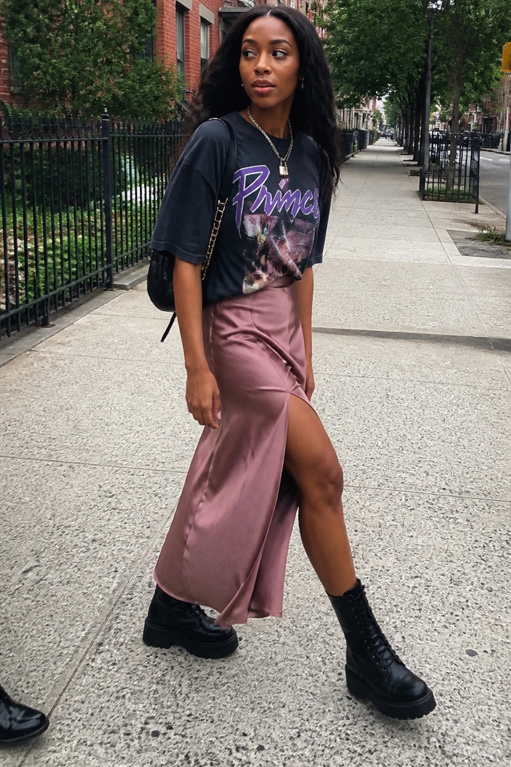 Woman wearing an oversized black Prince graphic t-shirt tucked into a dusty pink satin midi skirt with a high front slit, paired with black combat boots and a black chain-strap shoulder bag, plus layered necklaces.