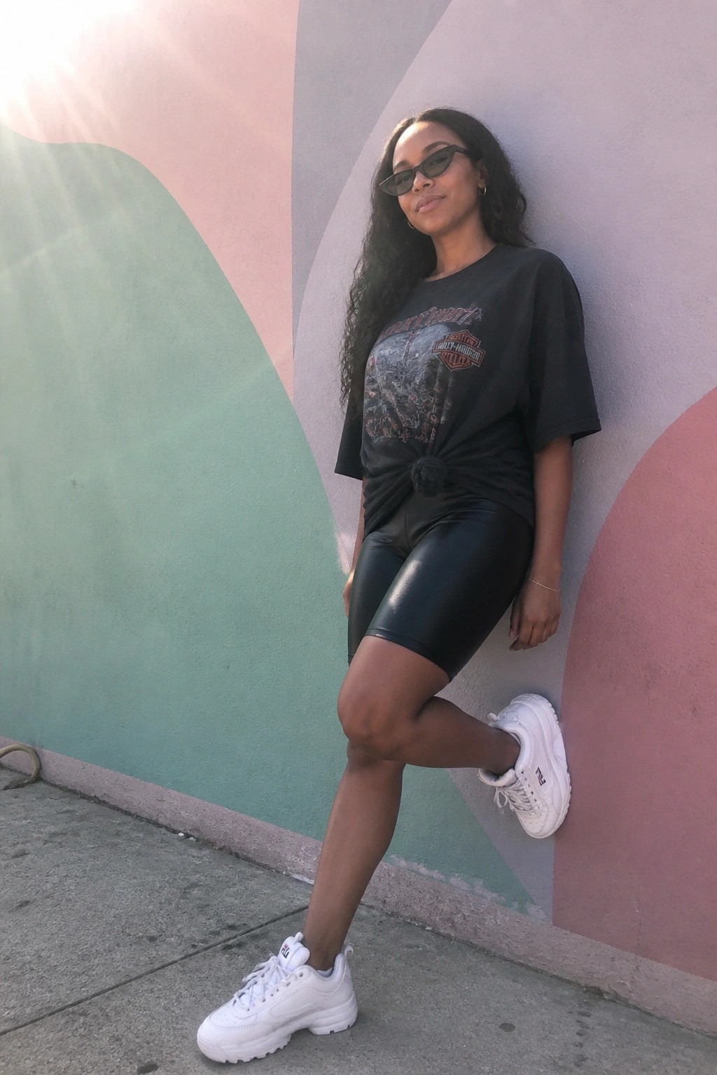 Woman leaning against colorful mural in black Harley tee, leather shorts, and white sneakers