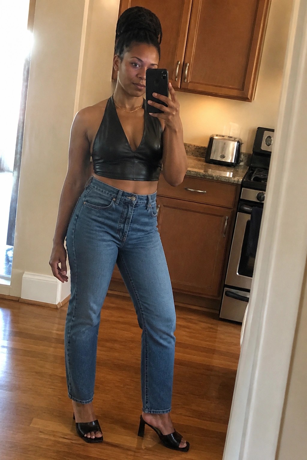 Woman in black halter crop top and blue jeans taking mirror selfie in kitchen