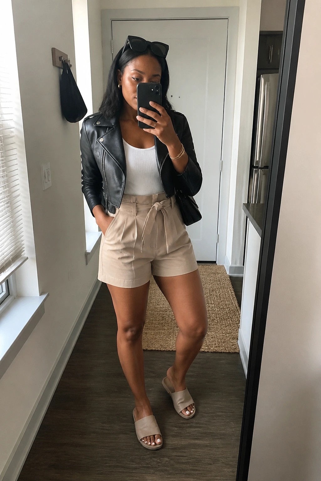 Woman in mirror selfie wearing black leather moto jacket over white ribbed tank top with beige high waisted tie belt shorts and matching slide sandals plus gold jewelry and sunglasses on head.