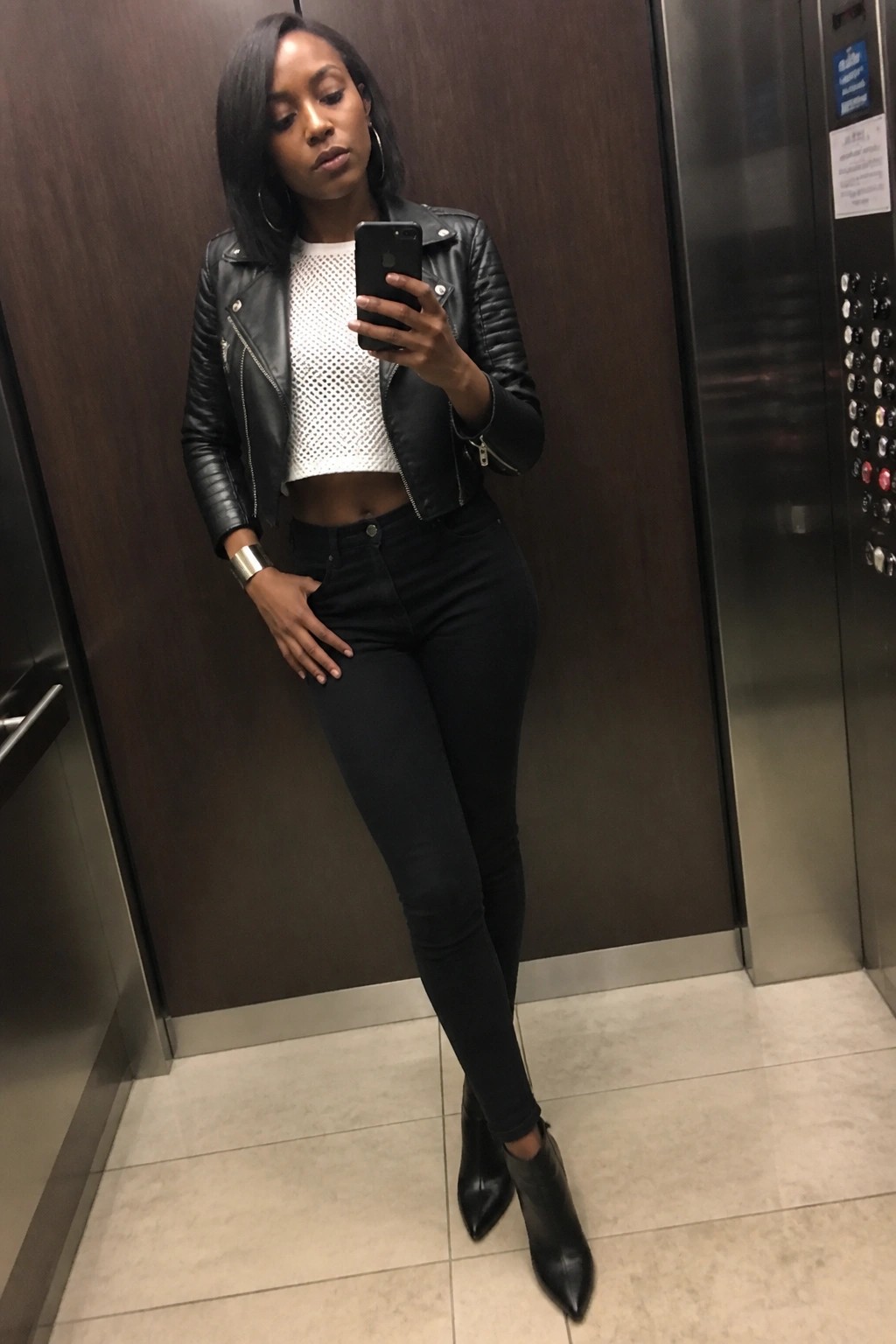 Woman in black leather jacket and jeans taking selfie in elevator.